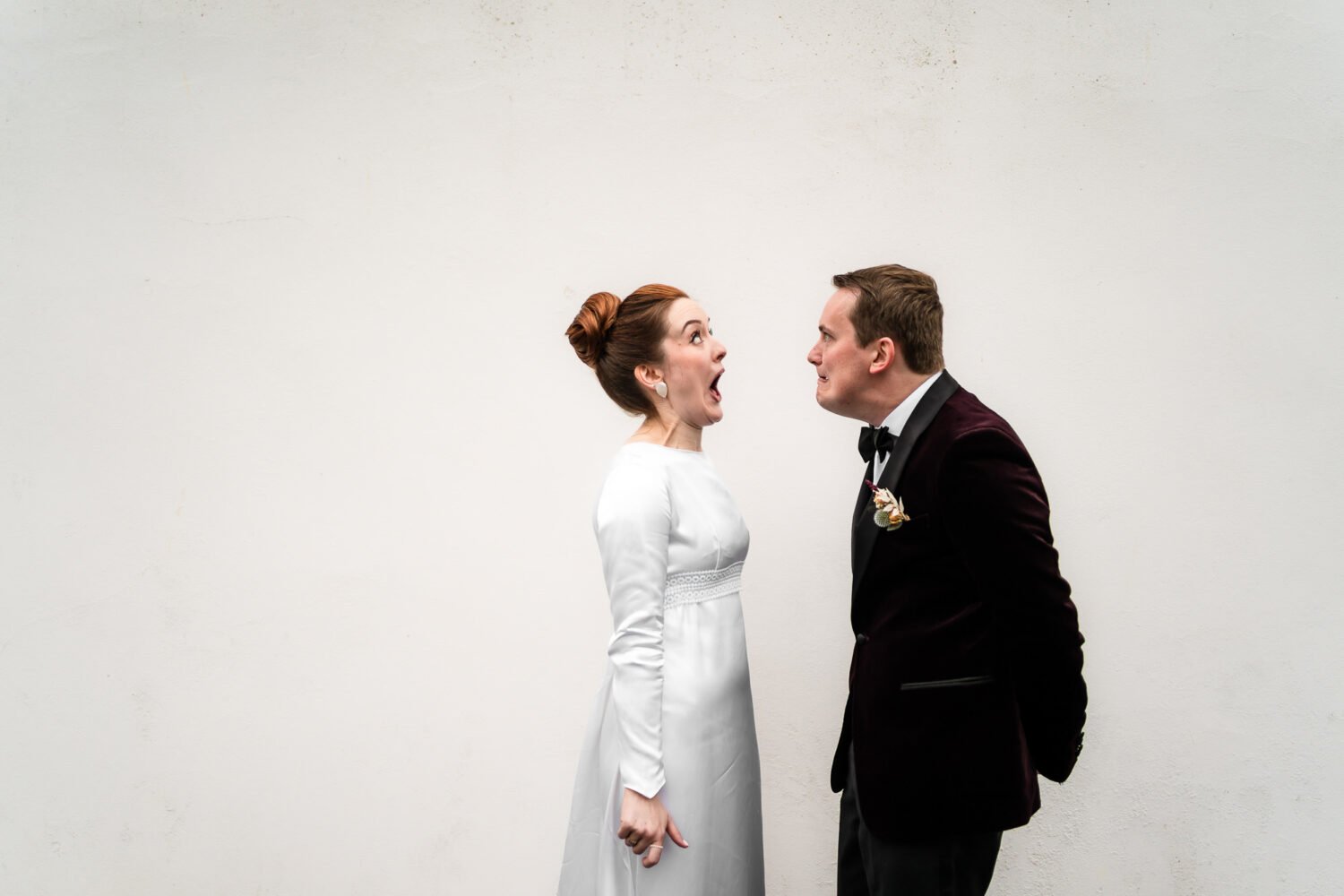 Bride and Groom endearingly pull funny faces at each other whilst stood in front of a white wall. How much does wedding photography cost?